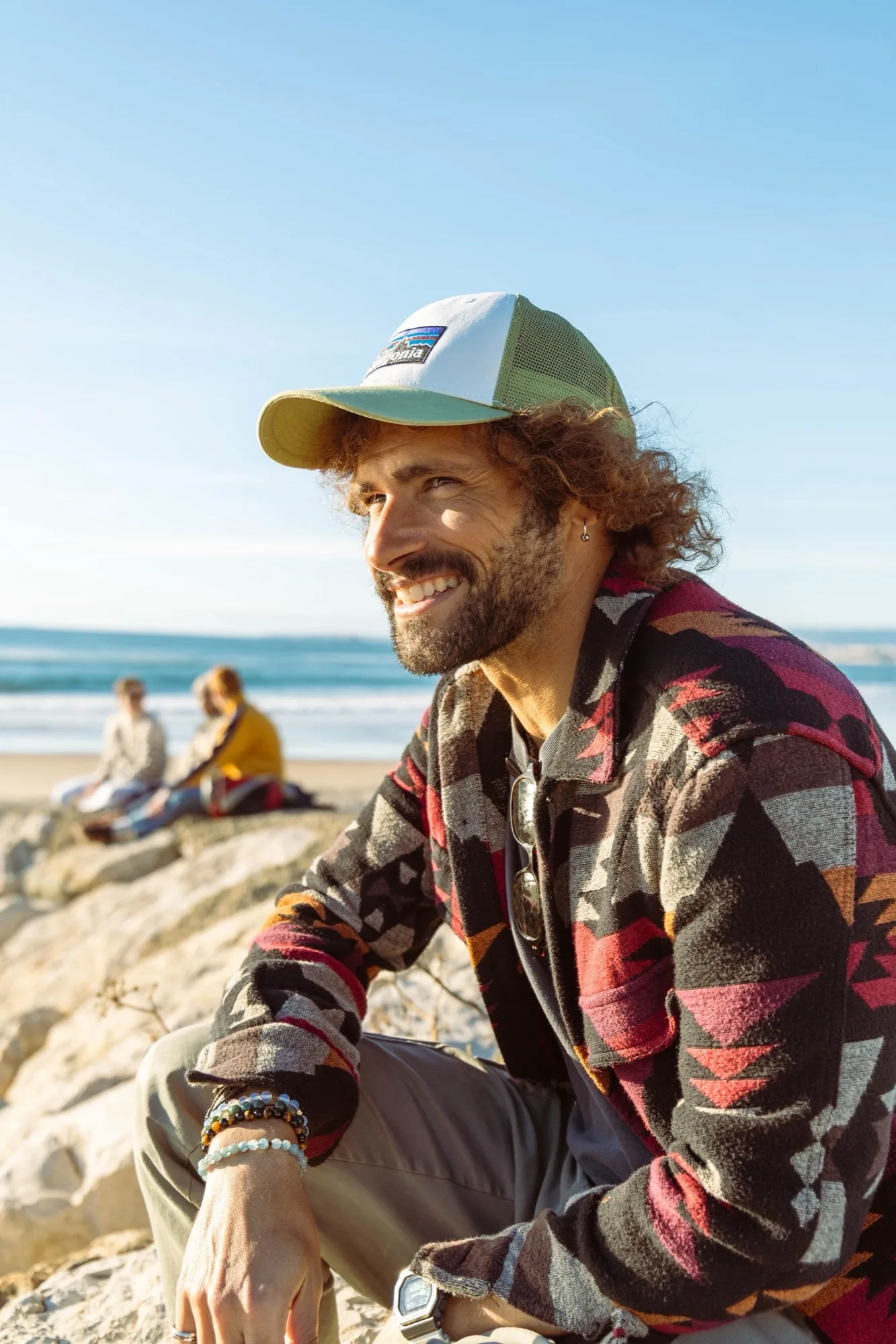 The photographer smiling on the rocks by the beach, in a patterned jacket and cap